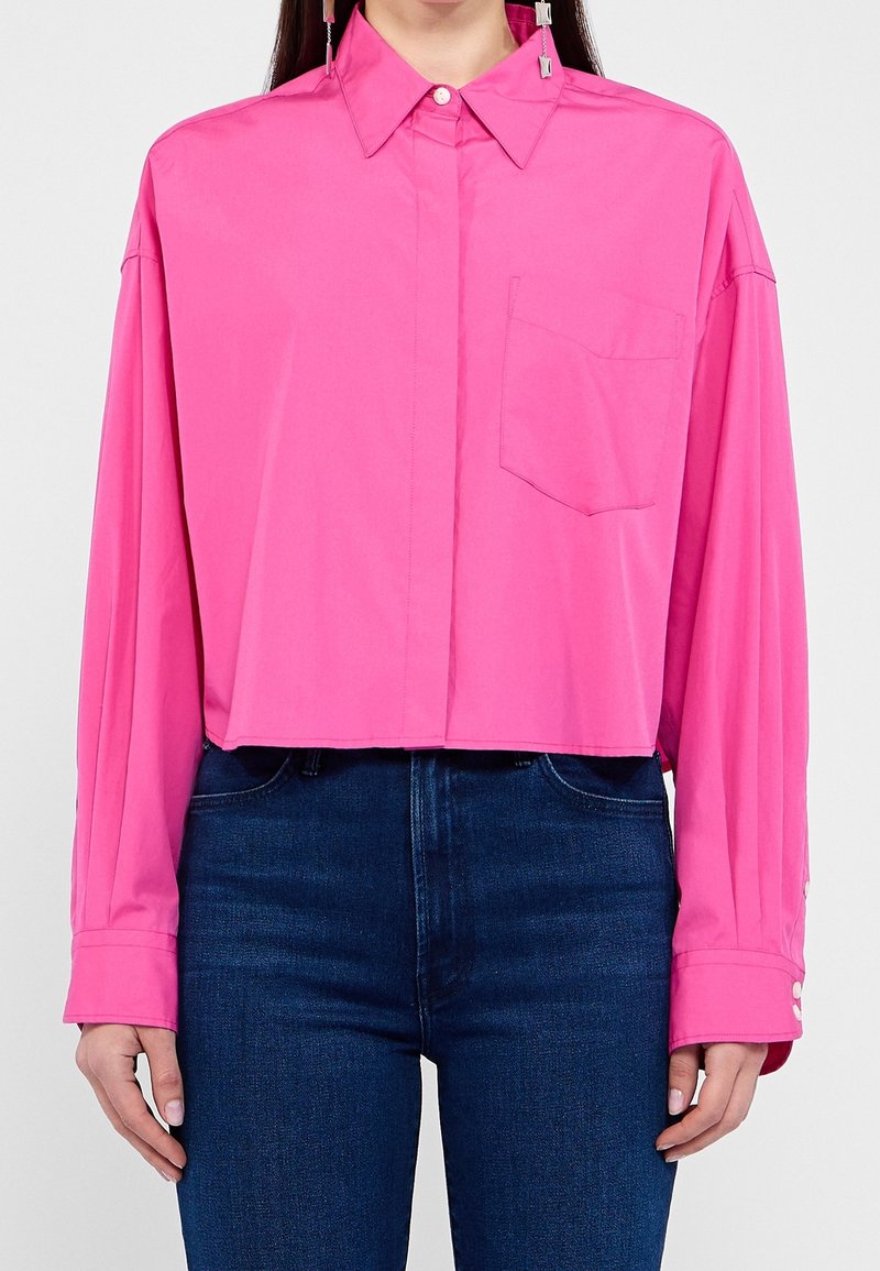 Personne portant un crop top rose vif à manches longues avec une poche devant et un jean taille haute bleu foncé, debout devant un fond uni.