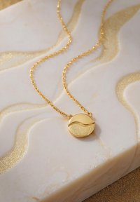 Guldkedja med en cirkulär berlock som har en texturerad vågdesign, mot en marmorbakgrund med en blank yta.