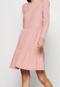 Abito rosa a costine con maniche lunghe, scollatura tonda e gonna a strati. Presenta spalle arricciate e un tessuto testurizzato.