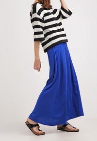 Haut oversize rayé noir et blanc avec des manches larges, associé à une jupe maxi fluide bleue et des sandales noires. Textures lisses tout au long.