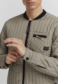 Giacca bomber trapuntata beige con rifiniture nere, chiusura frontale con zip e dettagli delle tasche. Presenta una superficie testurizzata e bottoni a pressione sui polsini.