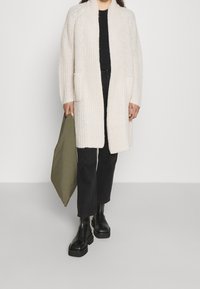 Krämfärgad stickad cardigan med öppen front, två fickor och ribbad textur, buren över en svart topp och matchad med svarta byxor och kraftiga stövlar.