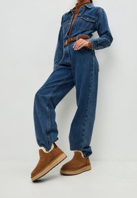 Persona che indossa una giacca di jeans blu e pantaloni con cintura marrone, dolcevita marrone e stivali slip-on foderati in pelliccia marrone, in piedi contro un muro bianco.