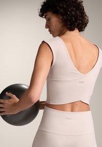 Világos rózsaszín sportfelső V-hát kialakítással, testhez simuló fazonnal és minimális varrásokkal, párosítva magas derekú, illeszkedő leggings-el, miközben fekete súlyt tart.