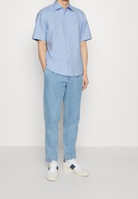 Hombre vestido con una camisa de boton corto de manga corta azul claro, jeans azul claro y zapatillas blancas con detalles en azul marino, de pie con las manos en los bolsillos.