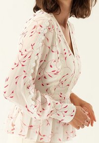 Blusa bianca trasparente con motivo floreale rosa, orlo in pizzo a scallop, maniche lunghe e fondo peplum. Tessuto texture con morbida caduta.