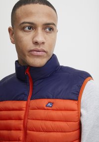 Polstret vest med en marineblå krave og orange krop. Har en høj hals, frontlynlås og et lille logo på brystet. Glat tekstur.