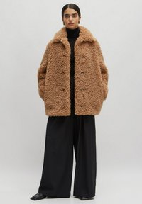 Flauscher, übergroßer brauner Mantel mit Schalkragen, ausgestattet mit drei Knöpfen, seitlichen Taschen und aus weichem, strukturiertem Stoff. Kombiniert mit schwarzen Hosen.