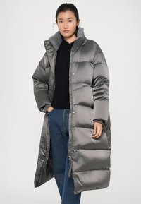 Grijze pufferjas met een licht glanzende textuur, lange lengte, drukknopen en een grijze faux bontkraag. Gecombineerd met donkerblauwe jeans.