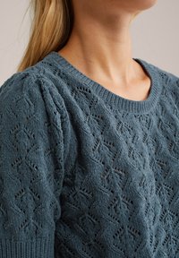 Maglione lavorato a maglia in blu teal con un motivo a trama aperta e testurizzata. Presenta un colletto rotondo e maniche arricciate per un tocco di dettaglio in più.