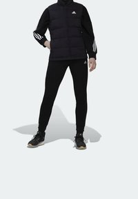 Svart vadderad väst med hög krage, svarta långärmade tröjor, tillsammans med svarta smala byxor. Logodetaljer på båda plaggen och sportiga sneakers.