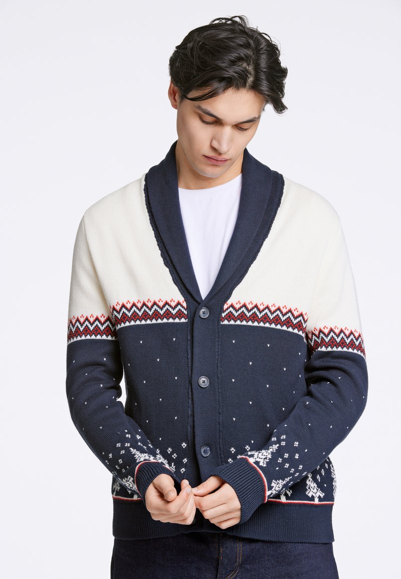 Marinblå och krämfärgad kofta med sjalkrage, knappframsida och festligt mönstrad ok. Har snöflinge-motiv och ribbade muddland.