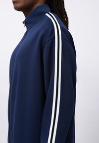 Navy zip-up jacket with high collar, featuring white and black striped accents on the sleeves. Smooth fabric and relaxed fit.