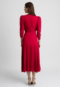 Czerwona sukienka z rękawami o długości trzech ćwierć, dopasowaną talią i rozkloszowanym dołem. Gładka tekstura, sięgająca kolan, zestawiona z beżowymi szpilkami.