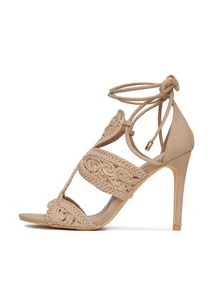 Beżowy sandał na wysokim obcasie wykonany z gładkiego materiału, z misternym haftowanym zdobieniem, otwartym noskiem i wiązaniem wokół kostki.