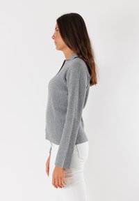 Maglione grigio a coste con colletto e maniche lunghe, caratterizzato da un design aderente e una texture sottile, abbinato a pantaloni bianchi attillati.