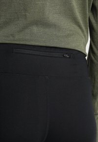 Černé legíny s hladkým povrchem a elastickým pasem, s malou zipovou kapsou vzadu. Vrchní část je textilní zelená košile s dlouhým rukávem.