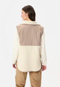Gilet beige et crème en tissu polaire doux avec un panneau arrière lisse et clair. Il présente un ourlet arrondi et un col.
