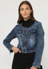 Chaqueta de denim con un diseño recortado, que presenta una cremallera frontal, dos bolsillos en el pecho y un lavado azul desvaído. Mangas largas con puños de botón.