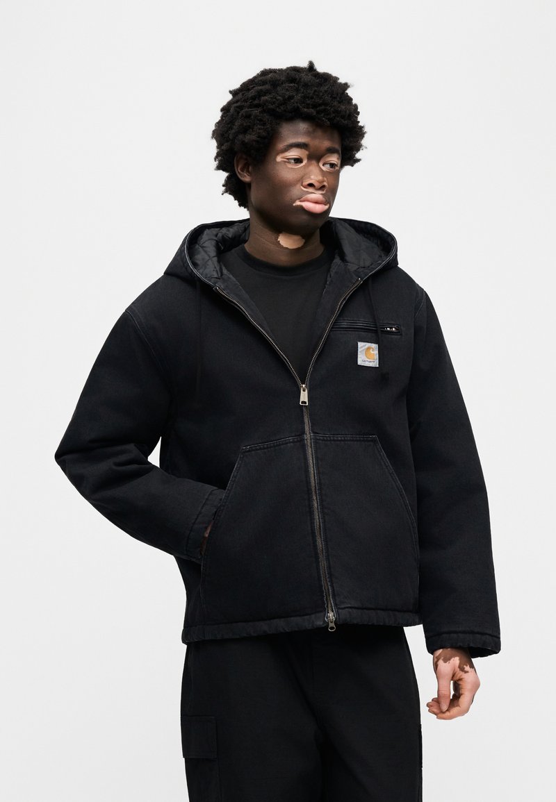 Veste noire à capuche avec fermeture éclair, poches avant et écusson logo. Le tissu est robuste avec une finition mate. Portée par-dessus une chemise noire.