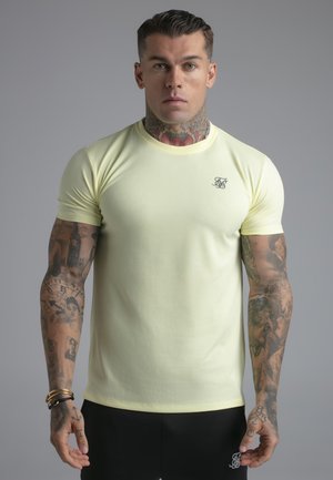 Mężczyzna z tatuażami na szyi i ramionach, ubrany w dopasowaną bladożółtą koszulkę i czarne spodnie, stoi tyłem do szarego tła.