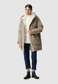 Giacca imbottita beige con cappuccio foderato in pile, tasche laterali e zip frontale. Indossata sopra un maglione crema e jeans blu, abbinata a stivali neri.