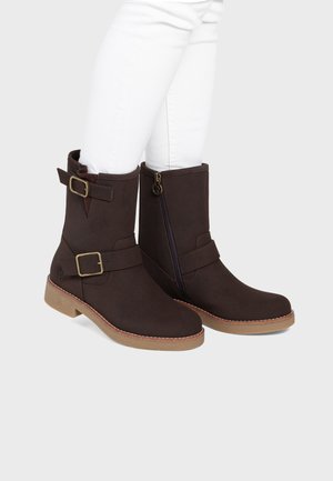 Bottines marron foncé avec fermeture éclair latérale et deux sangles à boucles portées avec un pantalon blanc sur fond clair.
