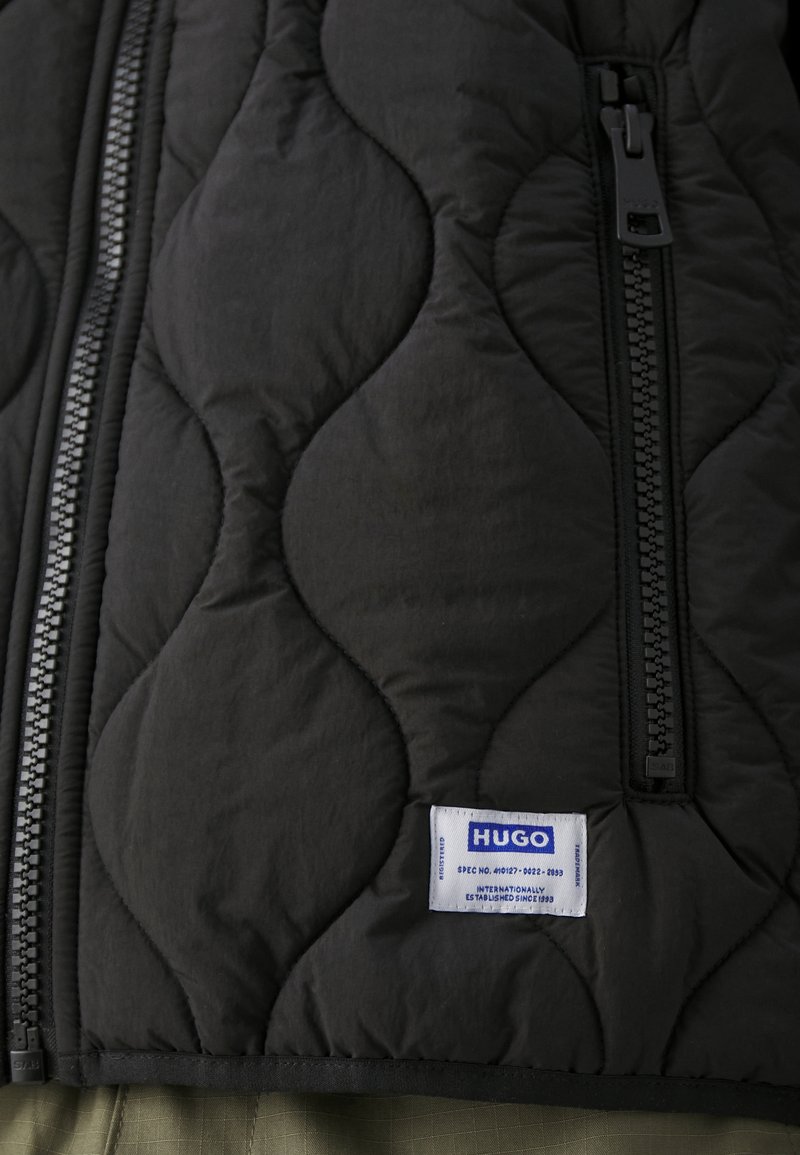 Czarna pikowana kamizelka z wyraźnym zamkiem błyskawicznym i prostokątną metką z niebieskim tekstem. Gładka struktura z falistym wzorem przeszyć.