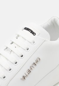 Hvit lær sneakers med flate hvite snøringer, metalløyer og preget logo detalj. Glatt tekstur med minimalistisk design.