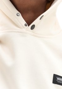 Cream-colored hooded sweatshirt made of soft fabric, featuring black snap buttons and a Vans logo tag stitched at the chest.