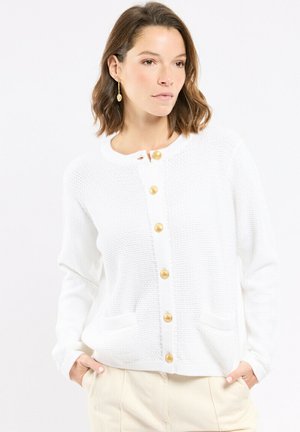 Femme aux cheveux bruns courts portant un cardigan blanc texturé avec des boutons dorés et un pantalon beige, les mains dans les poches, regardant vers la droite.