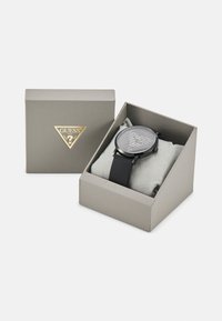Black watch with a round face, clear markings, and a silicone strap, presented in a light gray box with a triangular gold logo.