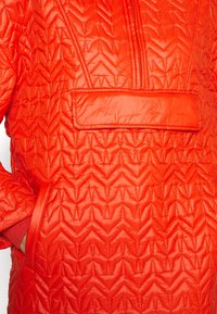 Czerwona pikowana kurtka z teksturowanym wzorem w zygzak, przednią kieszenią i wysokim kołnierzem. Wykonana z błyszczącego, lekkiego materiału.