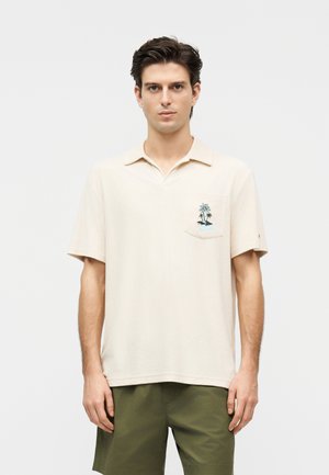 Jeune homme portant un polo à manches courtes beige avec une broderie de palmier sur la poche poitrine et un short vert, debout devant un fond blanc.
