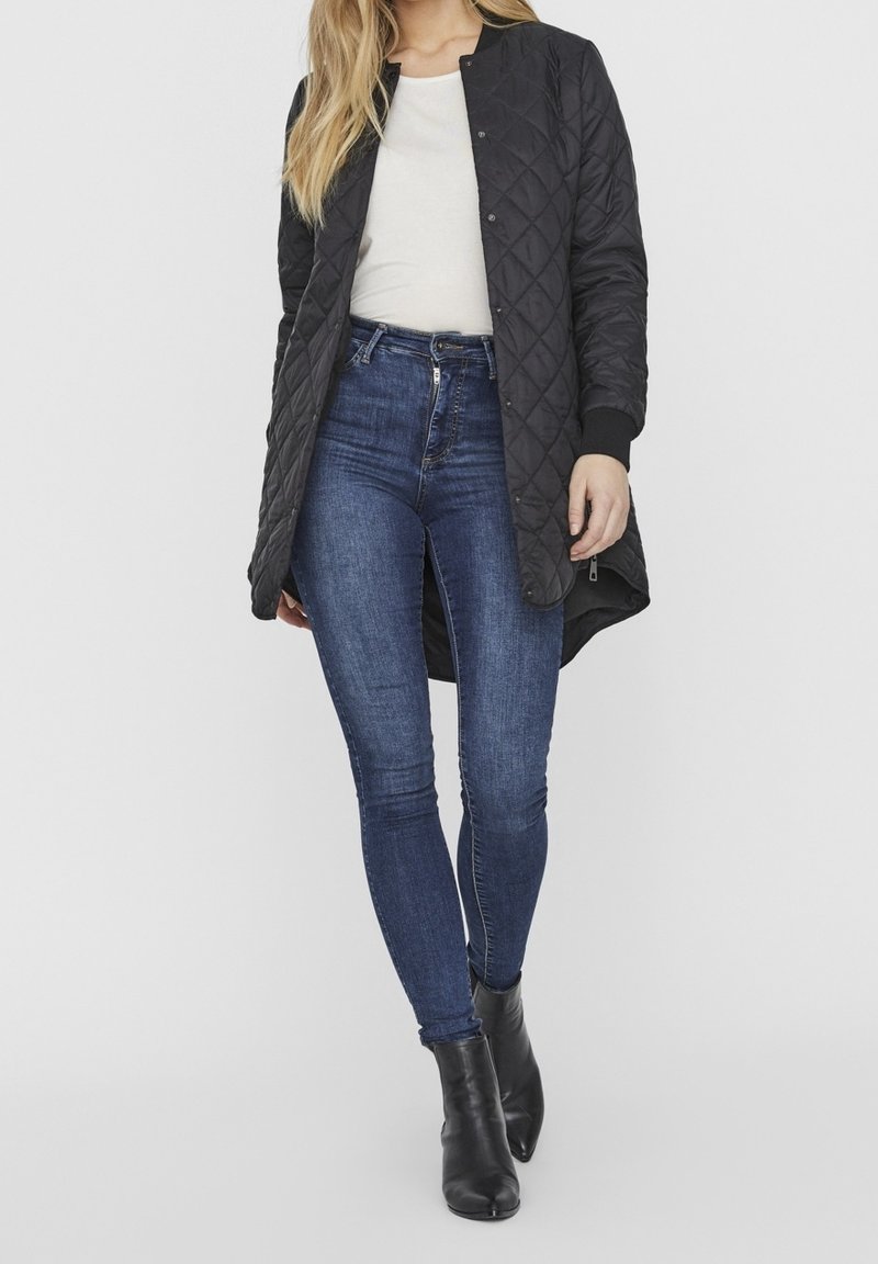 Giacca lunga nera trapuntata con scollatura rotonda, abbinata a jeans skinny blu scuro e stivaletti neri. Design semplice e funzionale.