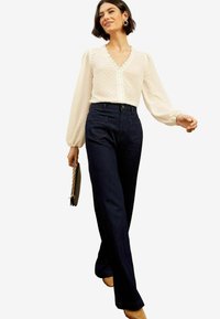 Blusa bianca con pois testurizzati e maniche a sbuffo, abbinata a pantaloni a piega in denim scuro a vita alta e una clutch nera.