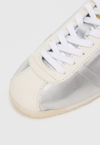 Adidași cu un superior din piele albă și accente metalice argintii. Prezintă șireturi albe, un vârf rotunjit și o talpă din cauciuc texturat.