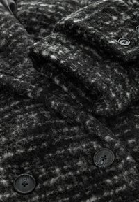Villane segu musta ja halli houndstooth mustris, millel on tekstureeritud pind ja mustad nupud. Näha on volditud varrukad.