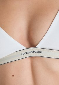 Close-up van een witte Calvin Klein bralette op de huid, met het merkteken op de elastische band onder de buste.