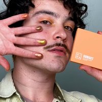 Palette de teintes chaudes dans un compact orange, avec du vernis à ongles métallisé doré et bronze sur les doigts, contre un fond clair.