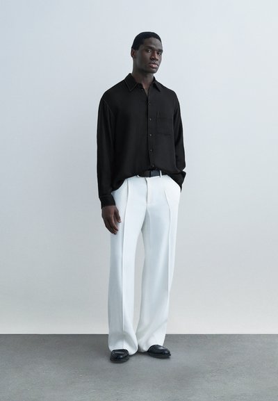 Άνδρας που στέκεται κοντά σε απλό τοίχο φορώντας φαρδιά μαύρο πουκάμισο με κουμπιά, λευκά φαρδιά παντελόνια, μαύρη ζώνη και μαύρα παπούτσια.