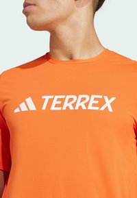 Orange athletic shirt featuring a large white "TERREX" logo. Made from a smooth, breathable fabric with short sleeves and a round neckline.