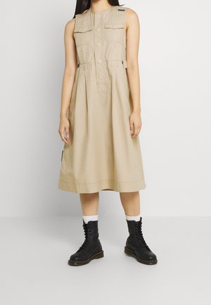 Robe midi beige sans manches avec boutons sur le devant, poches poitrine, fermetures éclair latérales, portée avec des chaussettes blanches et des bottes noires à lacets.
