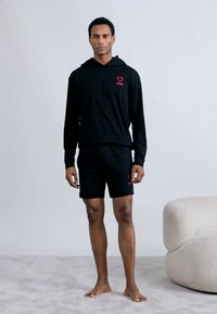 Μαύρο σετ φούτερ με κουκούλα και σορτς από βαμβακερ ό ύφασμα· κόκκινη καρδιά λογότυπο στο στήθος· το μοντέλο στέκεται ξυπόλητο σε ανοιχτό γκρι επιφάνεια.