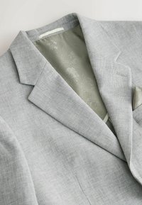 Hellgrauer Leinenblazer mit strukturierter Oberfläche, Revers mit Kerbe und einem gemusterten grünen Futter mit einem Einstecktuch.