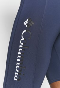 Námořnické modré sportovní šortky vyrobené z elastické látky, s velkým černým logem Columbia na boku a viditelnými švy.