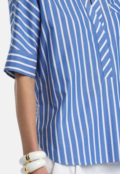 Bras portant une chemise à rayures verticales bleues et blanches avec manches retroussées, accessoirisé de bracelets blancs et dorés épais.