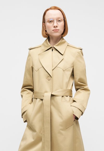 Γυναίκα με κοντά κόκκινα μαλλιά, φοράει γυαλιά και ένα μπεζ παλτό με ζώνη, με τα χέρια στις τσέπες, στέκεται μπροστά σε λευκό φόντο.