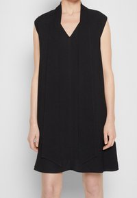 Svart ärmlös klänning med V-ringning och avslappnad passform, med vertikala veck och en utsvängd kjol. Tillverkad av mjukt, strukturerat tyg.