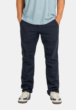 Man wearing navy blue pants, light blue shirt, and white sneakers with navy stripes, standing with hands in pockets against a plain background.